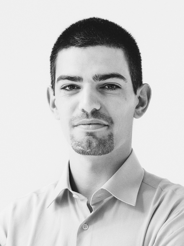 Black and white portrait of a young man with short hair and a goatee, wearing a light-colored button-up shirt, looking confidently at the camera.