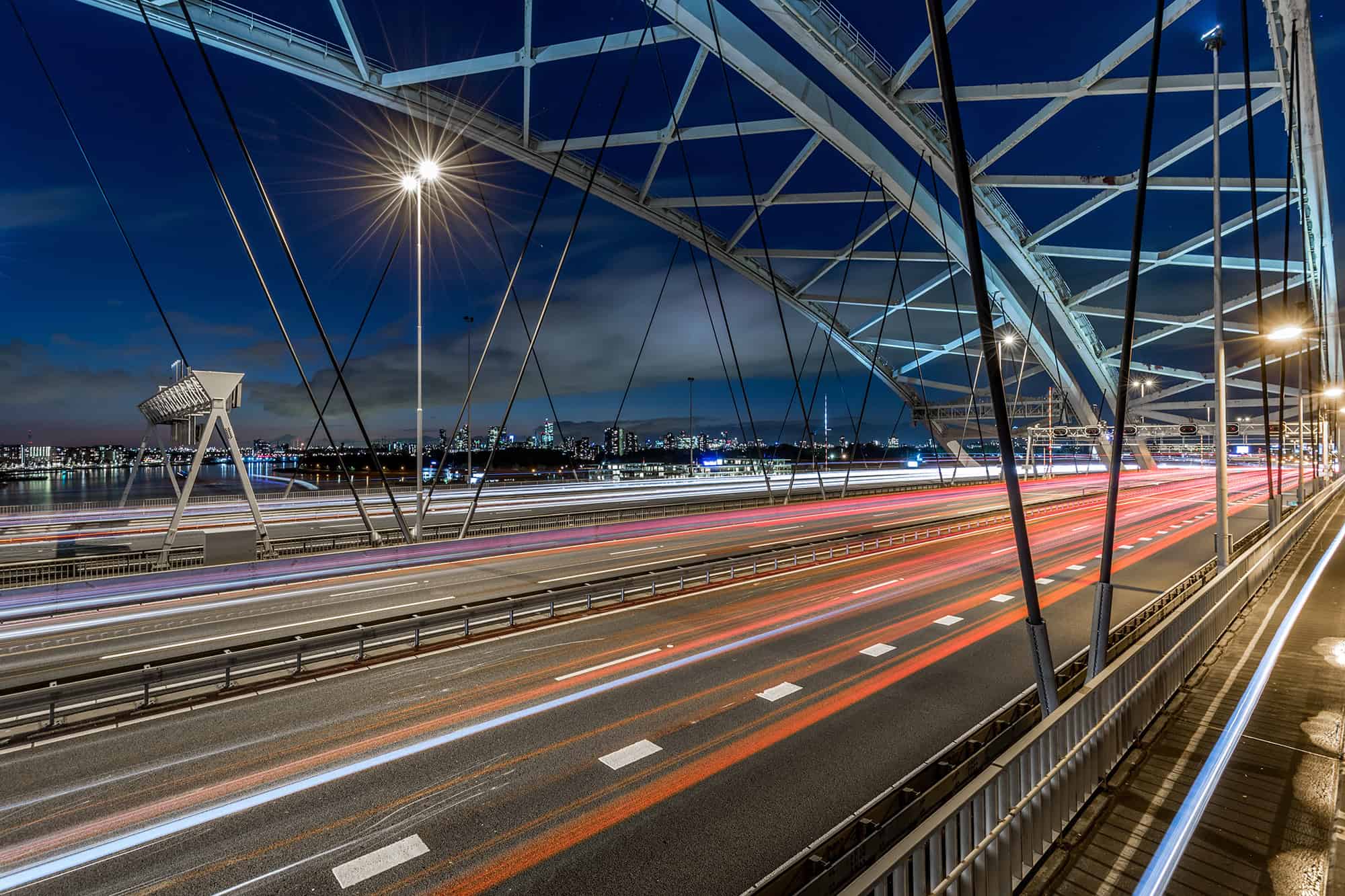 A long-exposure photo of a modern bridge at night shows light trails from traffic, illuminated streetlights, and a city skyline in the background under a partly cloudy sky.