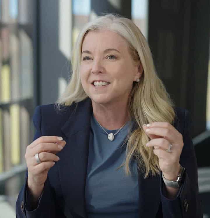 A woman with long blonde hair, wearing a navy blazer and blue top, gestures with her hands while speaking in a well-lit indoor setting.