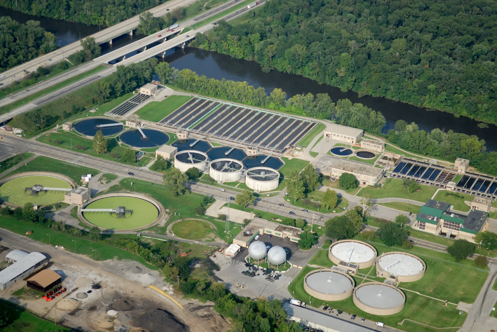 Aerial view of a wastewater treatment plant with multiple circular and rectangular tanks, adjacent to a river and surrounded by greenery, roads, and buildings.