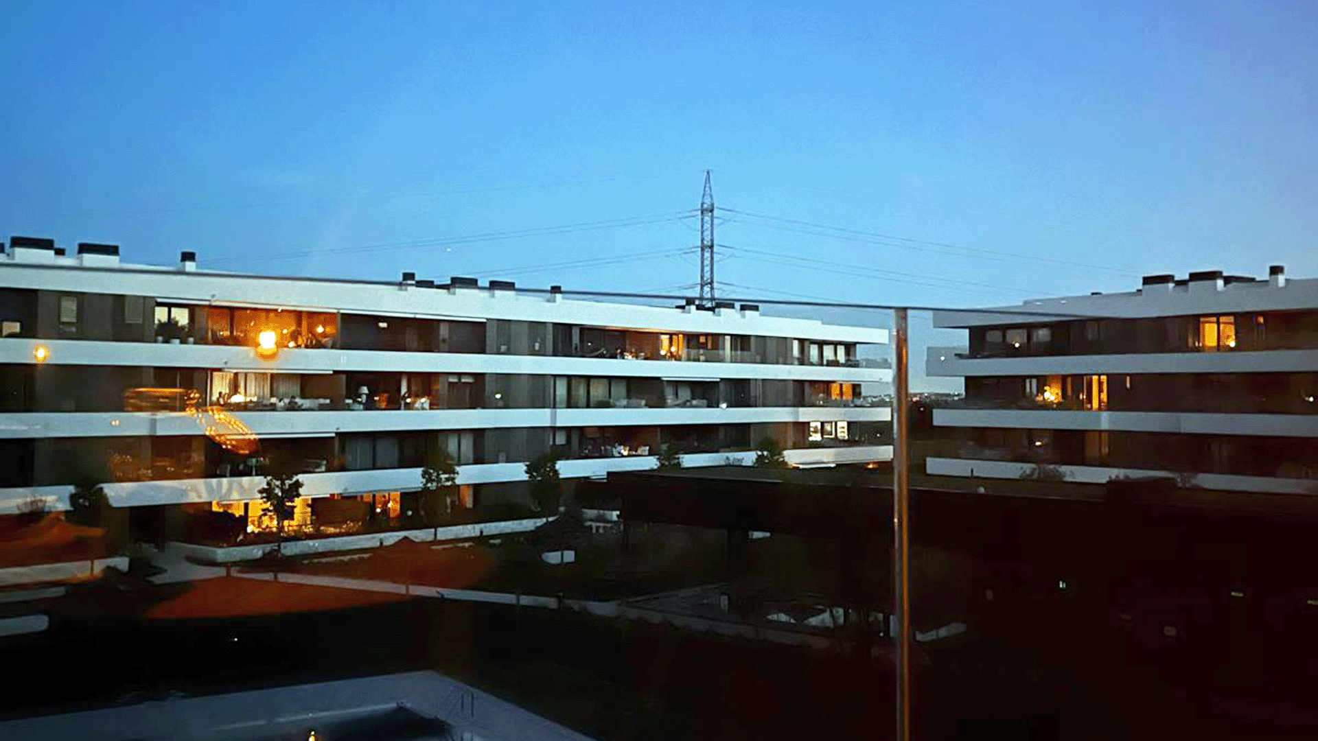 View of modern apartment buildings at dusk, their illuminated windows hinting at Stories inside, while a power line tower stands in the background—a scene echoing memories of the Iberian Blackout when all the lights went out.