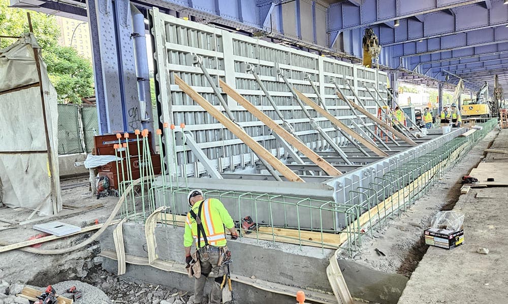 Construction workers in NYC reinforce and inspect a large concrete form under a bridge, with steel supports and temporary wooden braces visible, ensuring climate resilience in modern infrastructure.