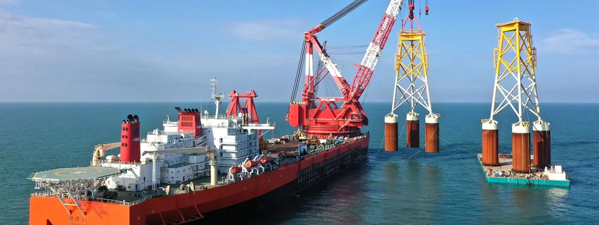 A large offshore construction vessel equipped with cranes installs two tower structures in the ocean, with a smaller support vessel nearby.
