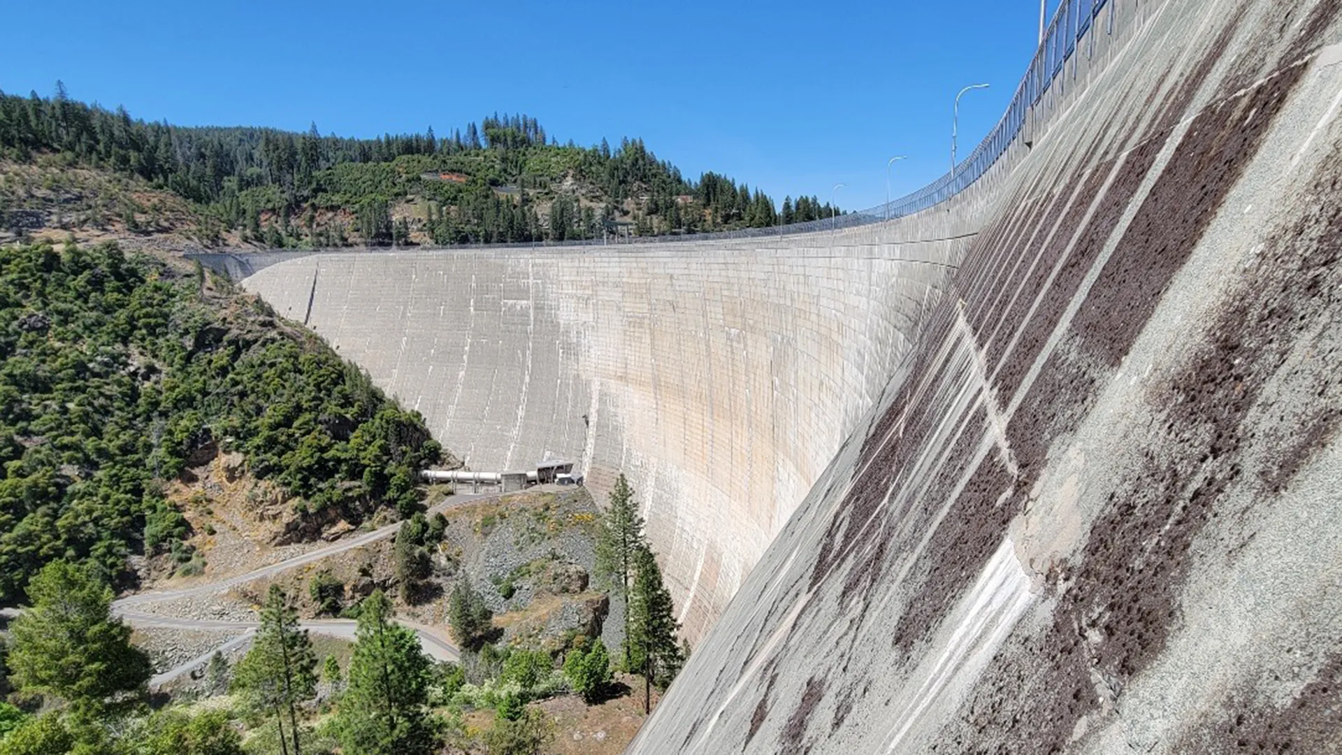 Large concrete dam with a curved face, managed by Yuba Water Agency, bordered by forested hills and a blue sky, with a road and small structures at the base ensuring public safety.