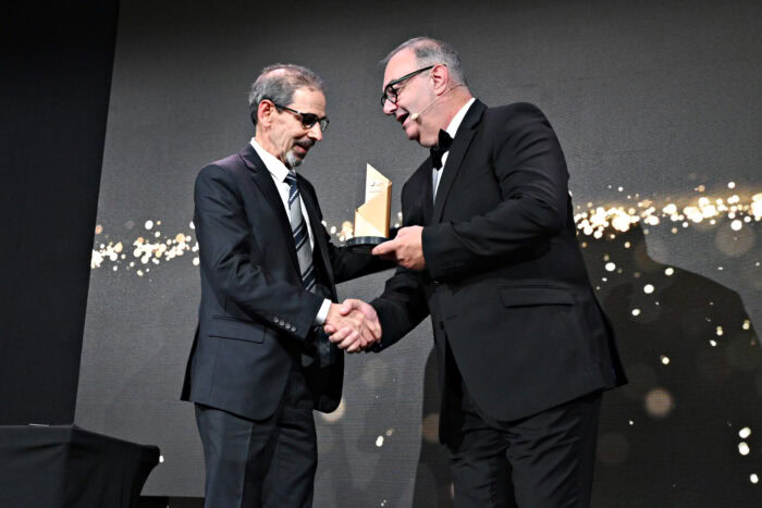 Two men in suits shake hands as one presents an award to the other on a stage with a black and gold backdrop.