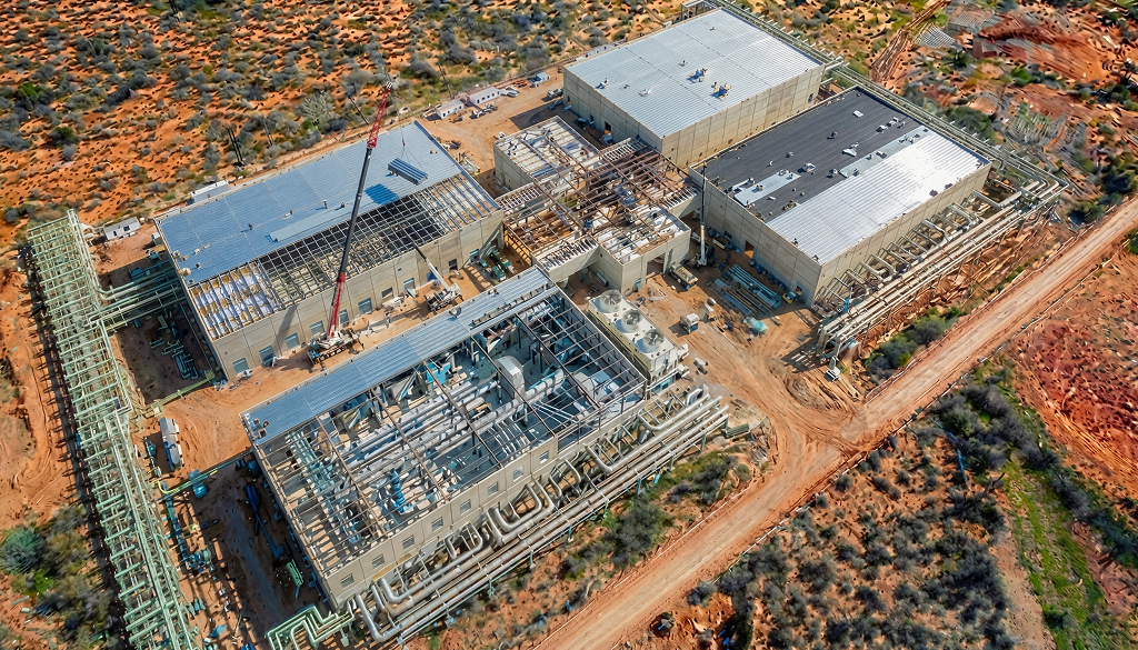 Aerial view of a data center delivery under construction in a desert area, featuring multiple large buildings, cranes, and extensive piping infrastructure.