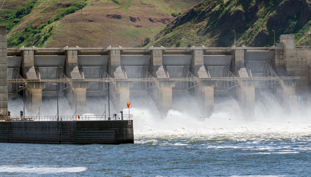 A large concrete dam, part of smarter infrastructure projects, releases water into a river, with mist rising at the base and green hills visible in the background.