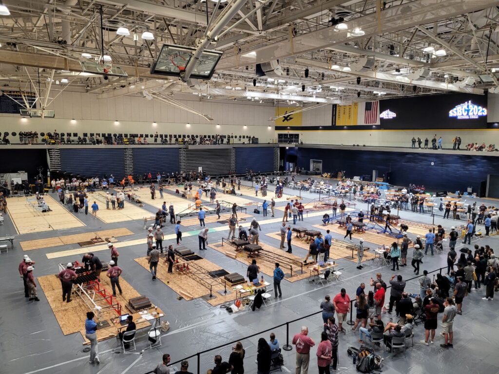 Large indoor gym hosting a robotics or engineering competition, where multiple teams work at tables as spectators watch—an exciting event that sparks innovation and fosters valuable career building for future engineers.