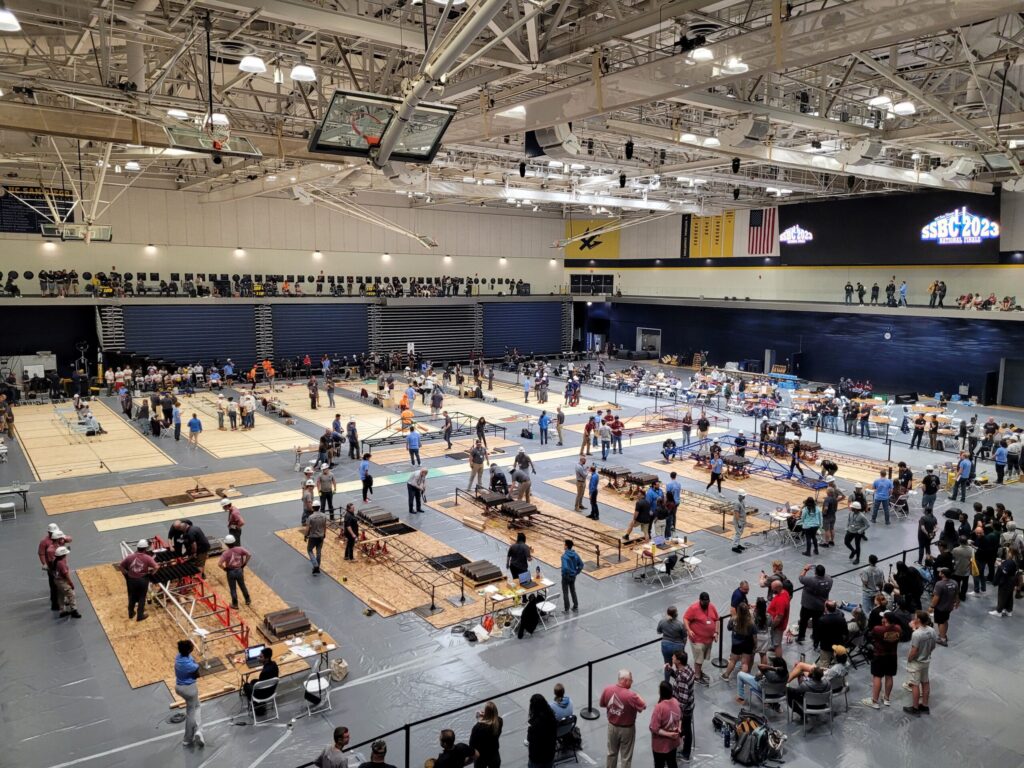 Large indoor gymnasium with multiple groups of people participating in a competitive event; audience members watch from the stands and sidelines.