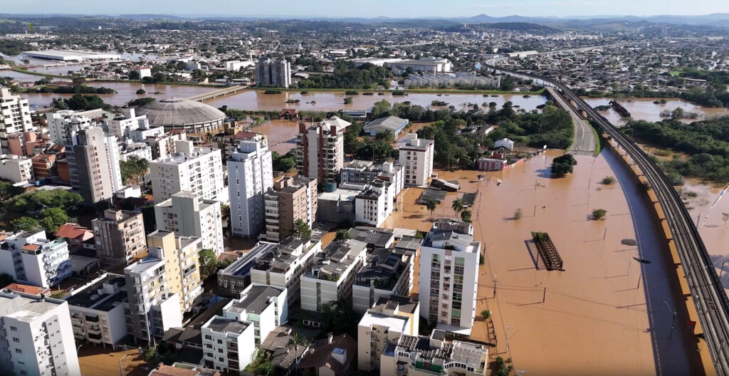 Aerial view of a city in Southern Brazil with flooded streets and buildings after the 2024 floods, brown water covering large areas, and a submerged bridge near a river as efforts for water supply restoration continue.