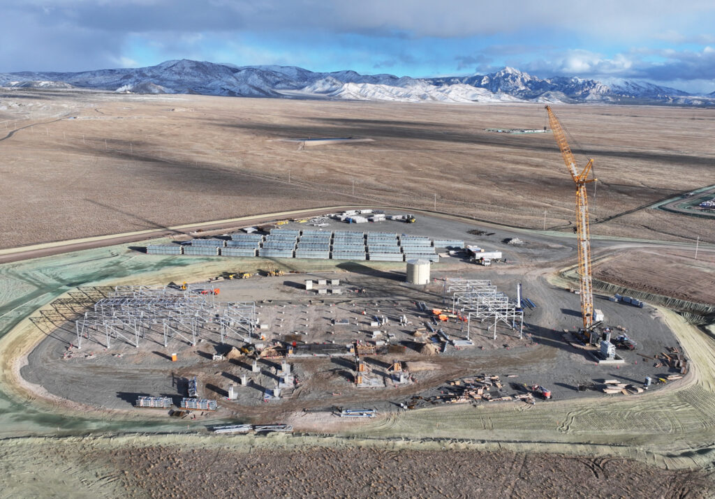 A construction site at Cape Station in a rural area shows steel framework, building materials, a large crane, and workers. Snow-capped mountains and open fields can be seen in the background.