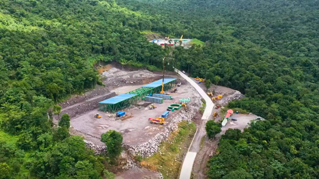 Aerial view of a Geothermal Project construction site with buildings, vehicles, and equipment, surrounded by dense green forest and a winding road in the Roseau Valley.