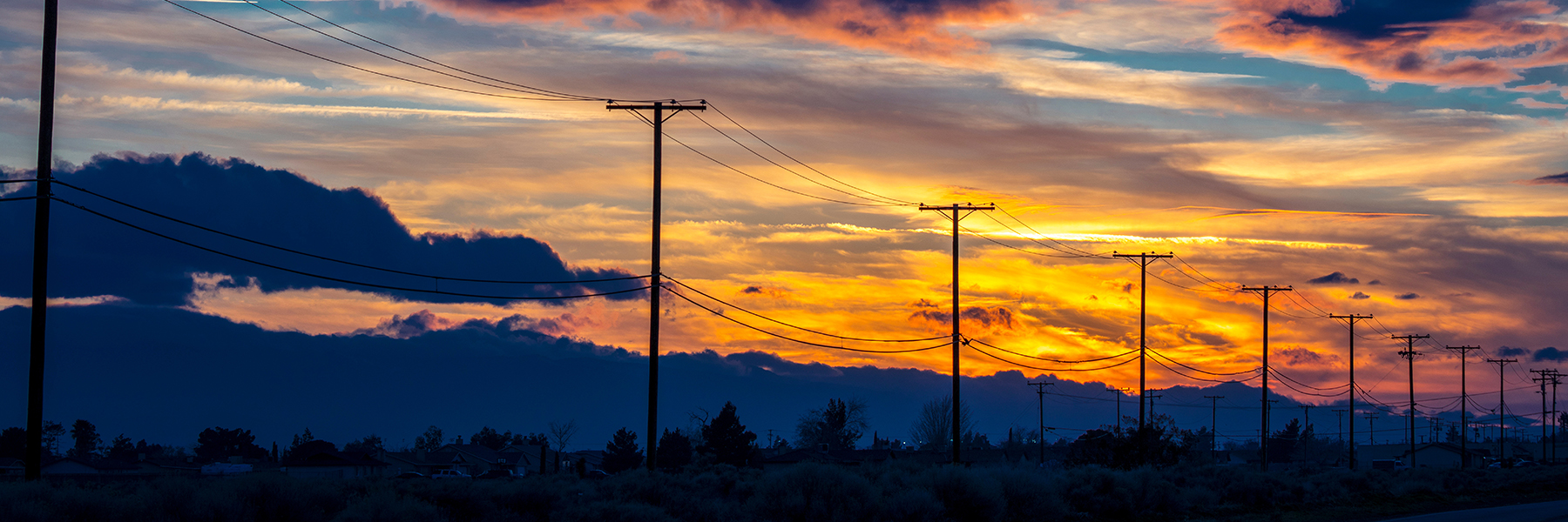 A sunset sky with vibrant orange and blue clouds, silhouetted by a row of electricity poles and wires, serving as a stunning backdrop for an electric distribution design demo.