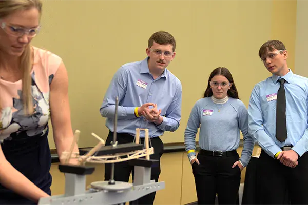 Three people, wearing safety glasses and formal attire, watch as a woman operates a small bridge model—an engaging scene that highlights hands-on learning for career development.