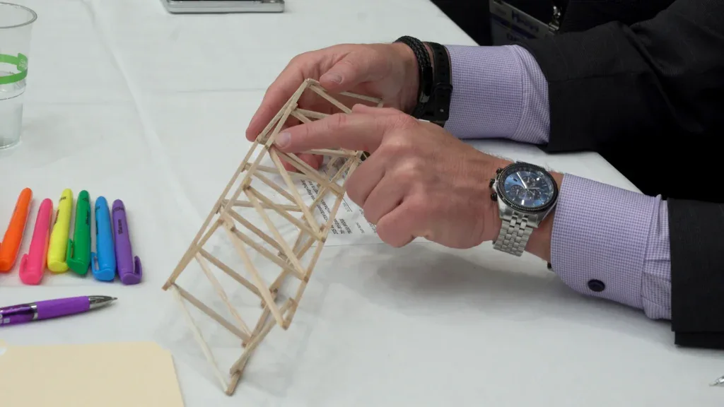A person in a suit points at a small bridge model made of wooden sticks