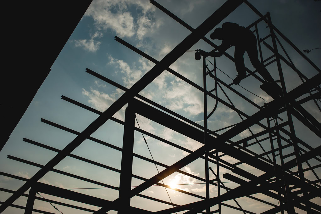 A worker is silhouetted against the sky while standing on scaffolding and working on a metal framework structure during construction, with the sun visible in the background.