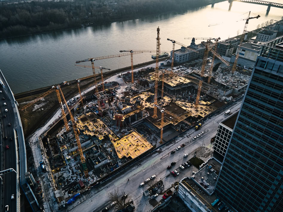 Aerial view of a large airport construction site with multiple cranes near a river, surrounded by roads and high-rise buildings.