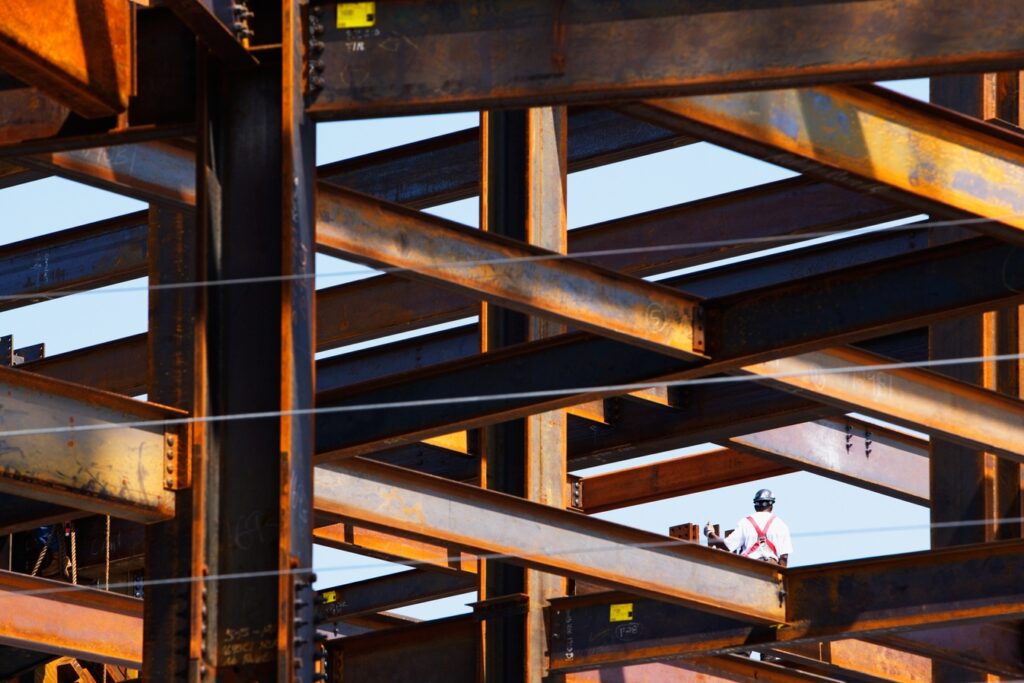 A construction worker wearing a safety harness stands among large, rust-colored steel beams at an construction site under a clear sky.