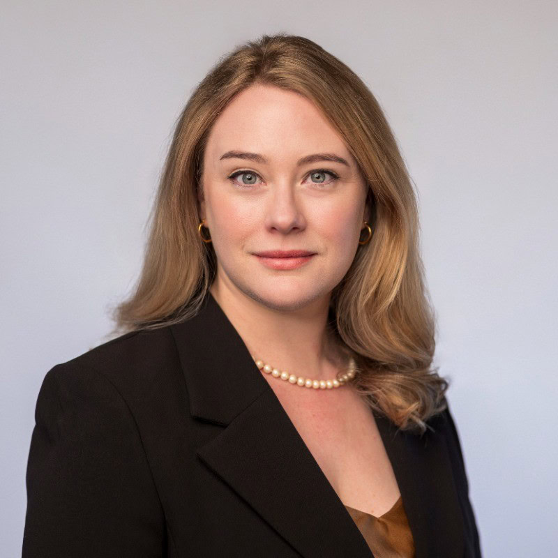 Kobieta z długimi do ramion blond włosami, ubrana w czarną marynarkę, brązową bluzkę, naszyjnik z pereł i złote kolczyki, pozuje pewnie na jednolitym jasnym tle – emanując dopracowanym profesjonalizmem zespołu Bentley.