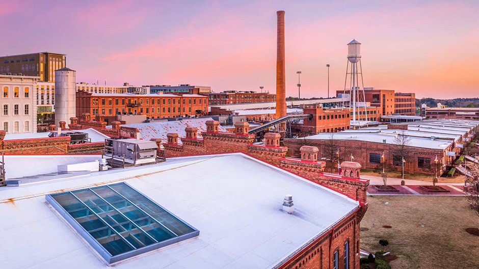 Brick industrial buildings with white roofs, a tall smokestack, and a water tower stand proudly under a pink and purple sunset sky—symbols of America’s infrastructure built to last in this vibrant urban area.