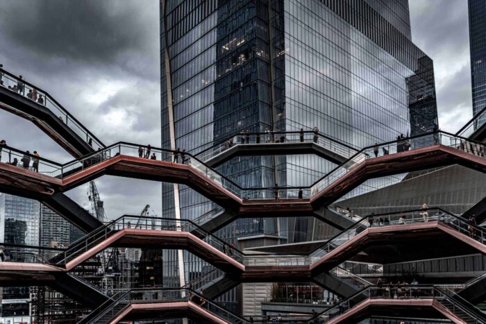 People walk on multiple levels of The Vessel, a honeycomb-like public structure in New York City, with glass skyscrapers and a cloudy sky in the background.