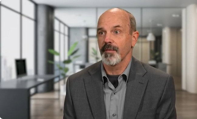 Partner Spotlight: A man with a gray beard and mustache, dressed in a gray suit and shirt, sits in a modern office with glass walls and plants in the background.