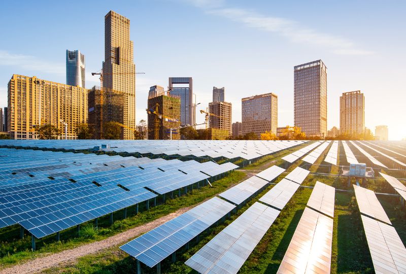 A field of solar panels glimmers under a clear sky, while in the background, a cityscape of tall buildings stands as a testament to Bentley Systems' Infrastructure Intelligence.