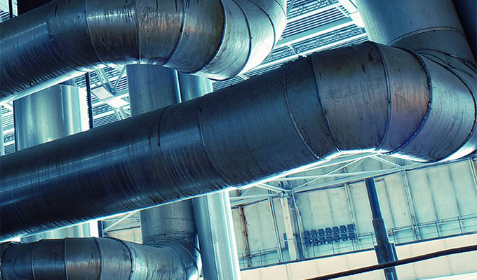 Large metal pipes and ducts in a modern industrial setting, with beams and structures visible in the background, illustrate an AssetWise infographic's precision detailing.