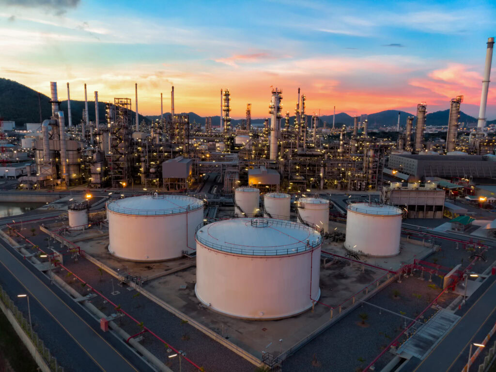 Aerial view of an industrial oil refinery, complete with Vessel storage tanks and towering processing units, captured at sunset. The scene, enhanced by AutoPIPE analysis, is encircled by roads and framed by a backdrop of rolling hills.