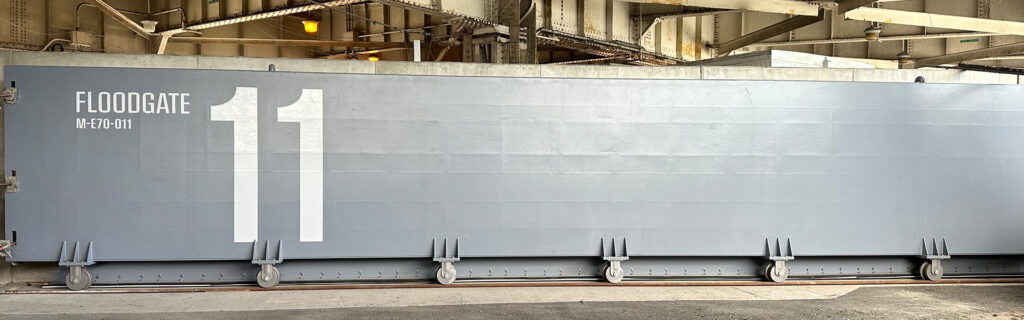Large gray Floodgate labeled "Floodgate 11 MF-F70-011" inside an industrial facility, with visible metal supports and overhead structural beams.
