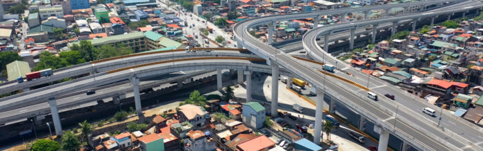 Aerial view of a complex multi-tiered highway interchange with vehicles, elevated above a densely built urban area comprising houses and buildings.