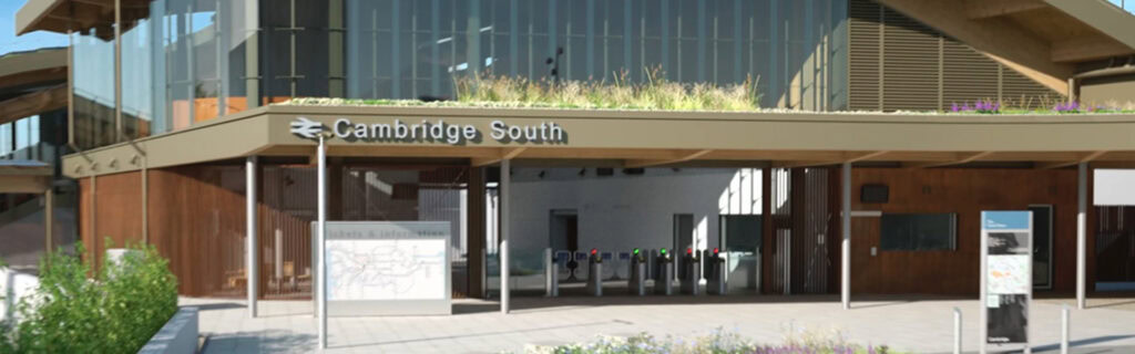 Cambridge South railway station entrance with signage, ticket barriers, and a map display. The station has a modern design with glass and wood elements and greenery on the roof.