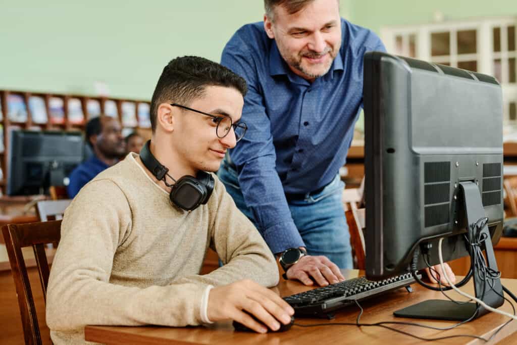 A middle-aged teacher assists a young male student with headphones who is using digital construction software on a computer in a classroom.