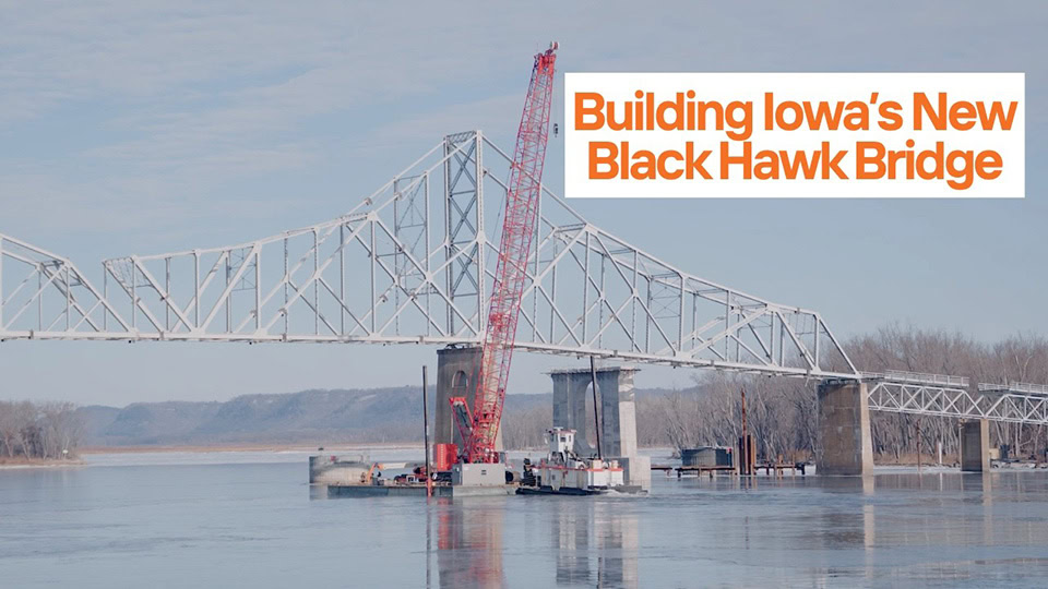 A construction crane and barge are positioned beneath the partially built Black Hawk Bridge— a steel structure that has been bridge built twice—spanning a wide river, with trees and hills in the background.