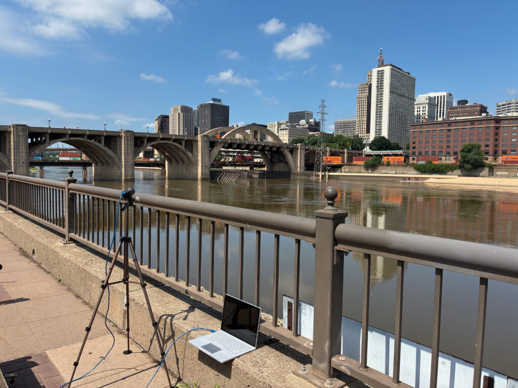 A laptop and tripod are set up on a riverside walkway, overlooking a bridge, train, and city buildings under a blue sky for the latest Roebling Labs Partner Spotlight.