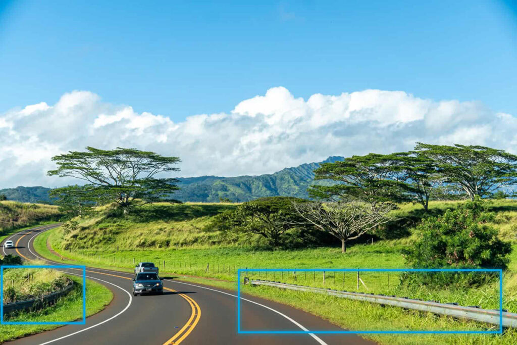 A car drives on a curved, two-lane road surrounded by grassy fields, trees, and distant mountains—showcasing impressive infrastructure engineering—while guardrails line the sides beneath a blue sky with clouds.