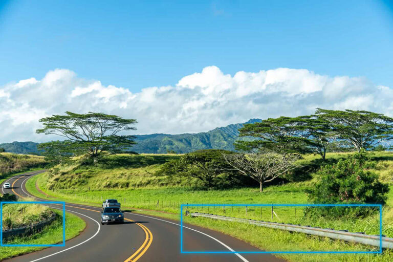A car drives on a curved, two-lane road surrounded by grassy fields, trees, and distant mountains—showcasing impressive infrastructure engineering—while guardrails line the sides beneath a blue sky with clouds.