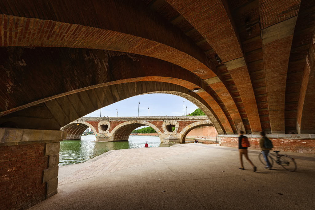 View from beneath a brick arch bridge overlooking a river, with two people walking and one person sitting by the water—capturing the quiet beauty of bridges and their tunnels framing peaceful riverside moments.