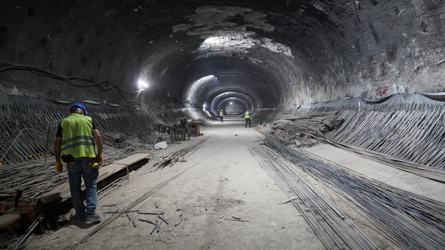 Construction workers in safety gear work inside a large, partially-built underground tunnel—an impressive example of tunnels construction—with exposed rebar and infrastructure materials along the walls and floor.