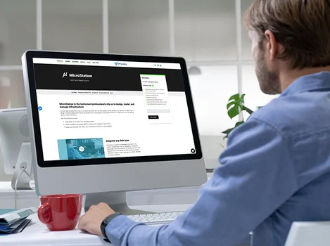 A person sits at a desk, looking at a computer monitor displaying a website called "MicroStation." A red mug is on the desk beside them, while the background features office decor. The screen reads “Make Your Mark with Design,” emphasizing creativity and innovation in every project.