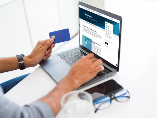 Person holding a credit card while using a laptop displaying a website, with a smartphone, glasses, and a pair of earphones on a white surface nearby. The setup captures the essence of modern infrastructure design for seamless online transactions.