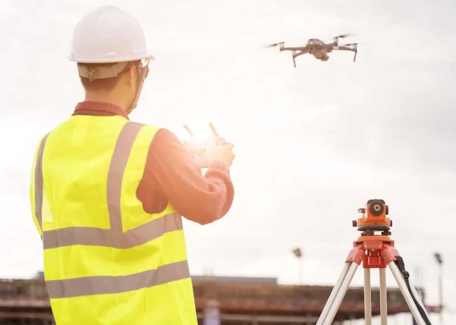 A person wearing a hard hat and high-visibility vest operates a drone with a remote control next to a tripod-mounted surveying instrument on a construction site, effectively integrating MicroStation software for precision in their mining projects.