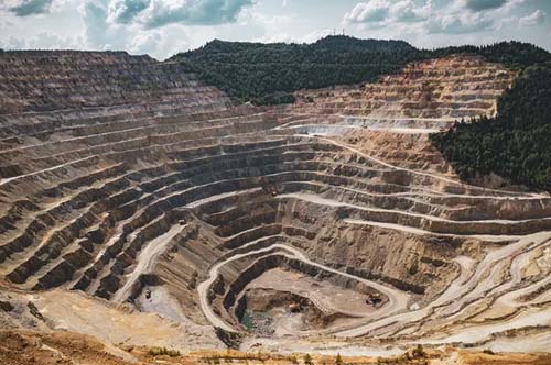 An open-pit mine with terraced layers descends into the earth, showcasing a MicroStation project, bordered by a forested area and cloudy sky in the background.