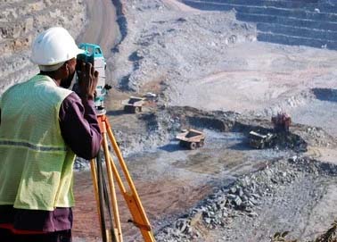 A person wearing a hard hat and high-visibility vest uses surveying equipment at a mining site, with large trucks and excavation visible in the background, all crucial elements for successful mining projects.