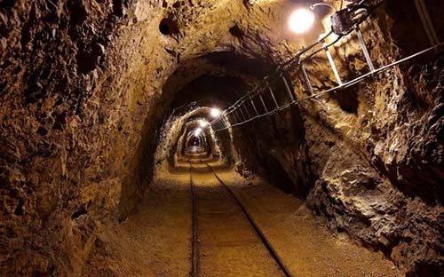 A dimly lit underground tunnel with tracks running along the ground and electrical wiring on the right side, features commonly seen in mining projects designed with MicroStation. The walls and ceiling are rough-hewn rock.