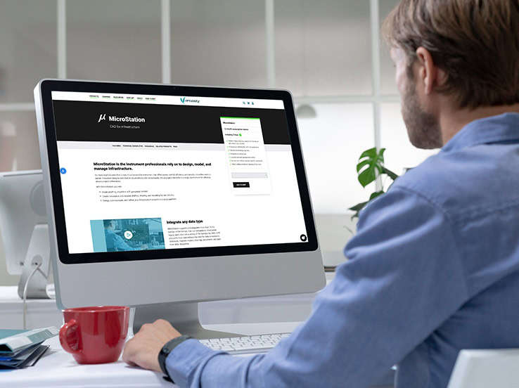 Man sitting at a desk views a microstation software page on his computer monitor, with a red mug beside him.