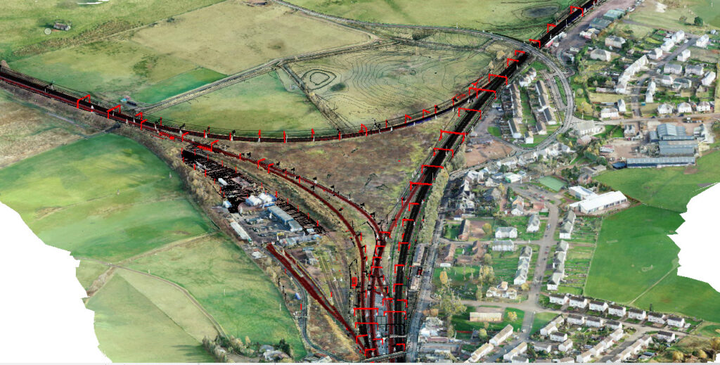 Aerial view of a railway junction intersecting green fields and residential areas, featuring transportation infrastructure and tracks marked in red.
