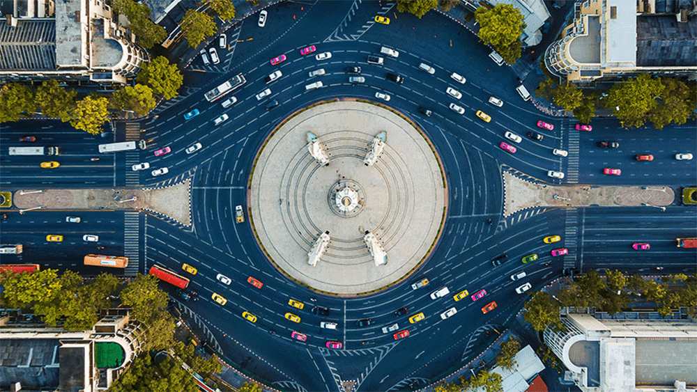 Aerial view of a large roundabout near the Barcelona Experience Centre, with statues at the center, multiple roads radiating outward, and various cars and buses circulating.