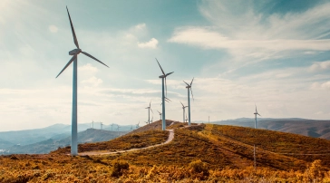 Wind turbines on a grassy hill generate renewable energy under a partly cloudy sky with distant mountains in the background.