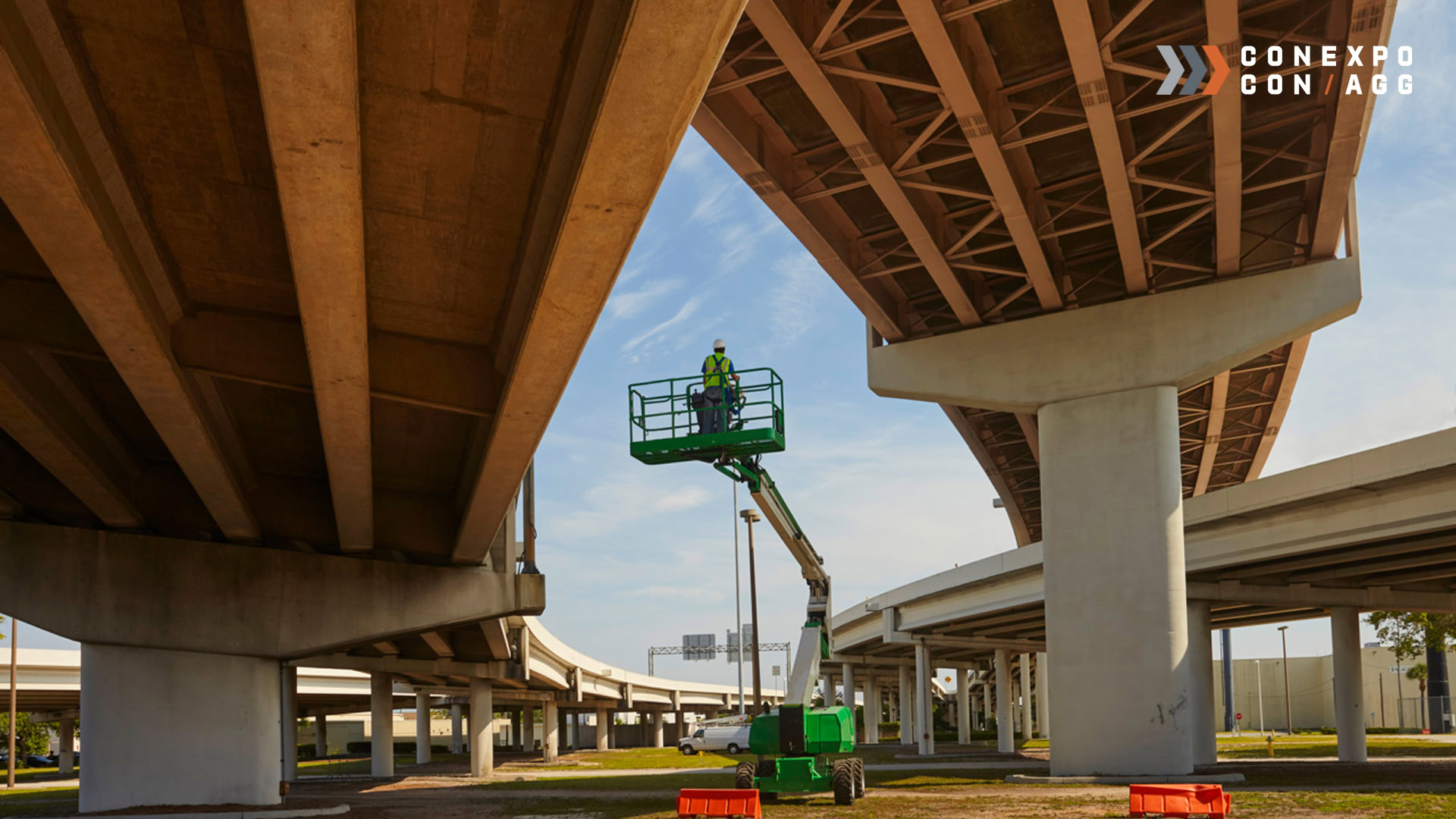 A worker in a safety vest stands on a green lift beneath a highway overpass, inspecting the structure. Multiple overpass lanes and concrete supports are visible.